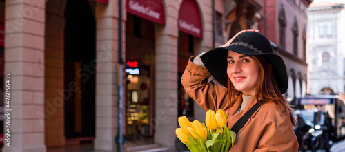 Beautiful young girl in a hat with a big bouquet of yellow tulips. Walks around the old town in European. Woman with flowers on the street. Copy space. Banner.