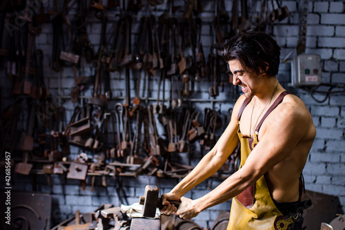 Authentic blacksmith man forges a metal product in dark indoors studio