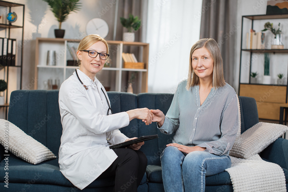 Front view of good-looking professional female Caucasian doctor in medical coat, consulting her senior patient and handshaking after sighning an insurance, during home visit. Front view of good-looking professional female Caucasian doctor in medical coat, consulting her senior patient and handshaking after sighning an insurance, during home visit.