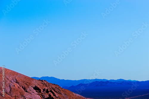 Barren reddish barren mountainside with purple and blue mountain ranges beyond under a hazy blue sky.