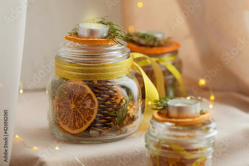 Hand-decorated candles with dry slices of citrus, cones, spruce twigs in glass cans on a light beige linen background. Eco jewelry concept for New Year and Christmas.