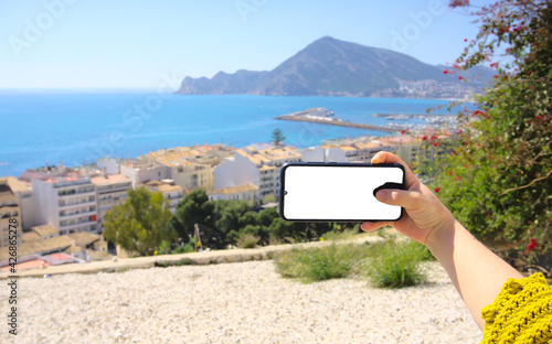 Hand holding mobile phone in front of the landscape of Altea, Alicante, Spain. White screen, copy space photo