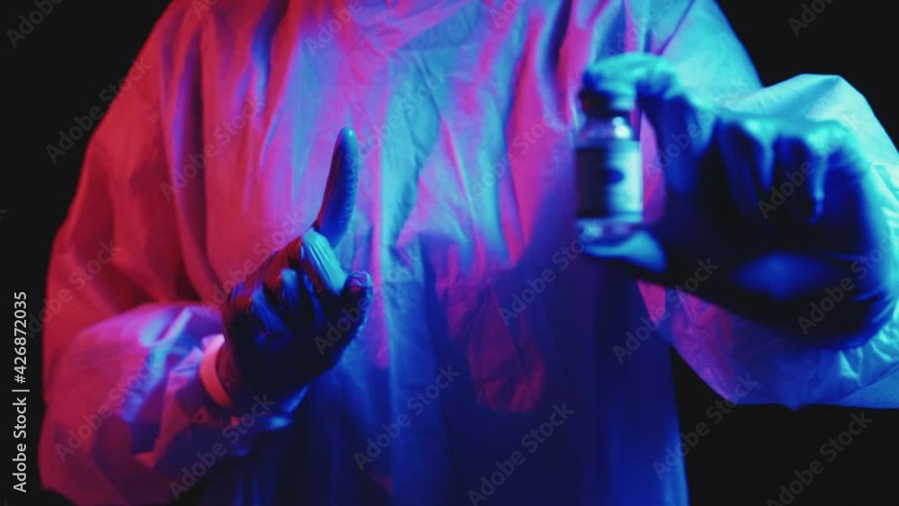 Vaccine risk. Covid-19 cure. Clinical trial. Doctor in blue ppe protective gloves warning with finger gesture showing dose vial in pink neon light on dark defocused background. Vaccine risk. Covid-19 cure. Clinical trial. Doctor in blue ppe protective gloves warning with finger gesture showing dose vial in pink neon light on dark defocused background.