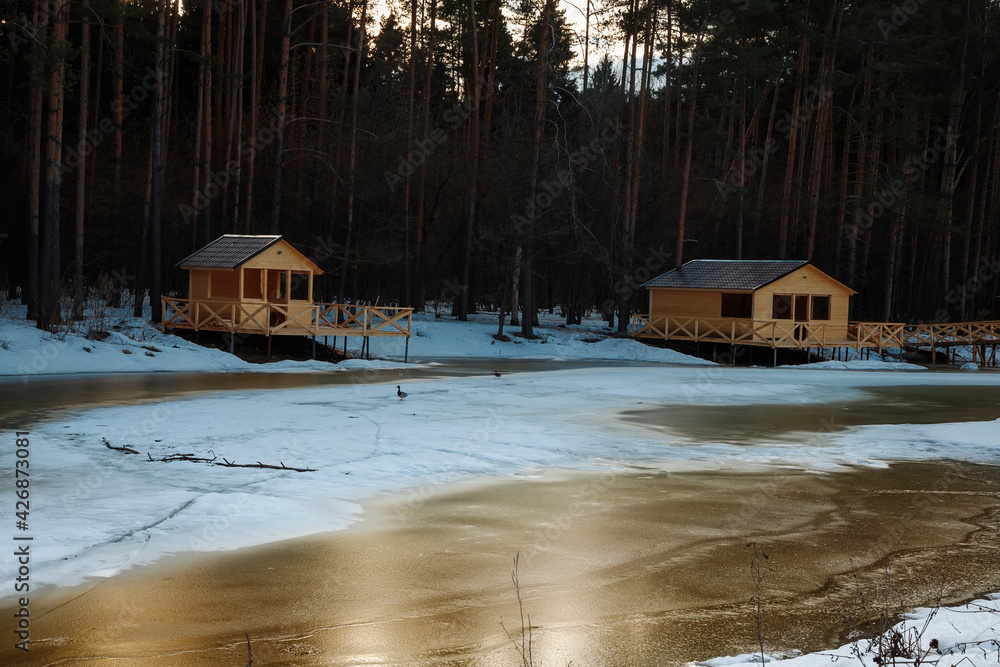 A wooden house on the shore of a frozen lake. Unfinished wooden bridge over the pond.