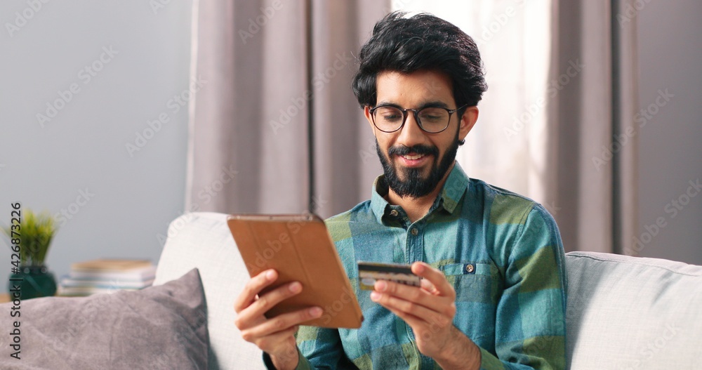 Portrait of Indian young happy bearded man in glasses shopping online buying on internet using tablet device making payment with credit card, e-commerce service, shopaholic buyer concept Portrait of Indian young happy bearded man in glasses shopping online buying on internet using tablet device making payment with credit card, e-commerce service, shopaholic buyer concept