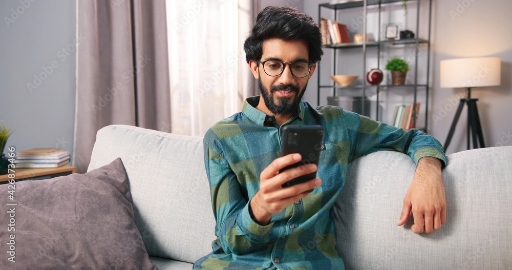 Close up of handsome young cheerful Hindu bearded guy sitting on couch at home in cozy living room and texting, browsing online on smartphone using social network app. Technology gadget Close up of handsome young cheerful Hindu bearded guy sitting on couch at home in cozy living room and texting, browsing online on smartphone using social network app. Technology gadget