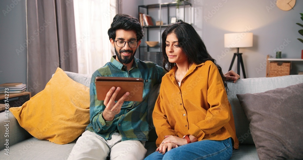 Hindu happy handsome young bearded husband browsing online on tablet surfing internet discussing with pretty wife some news on social network app, positive mood, family relations concept Hindu happy handsome young bearded husband browsing online on tablet surfing internet discussing with pretty wife some news on social network app, positive mood, family relations concept