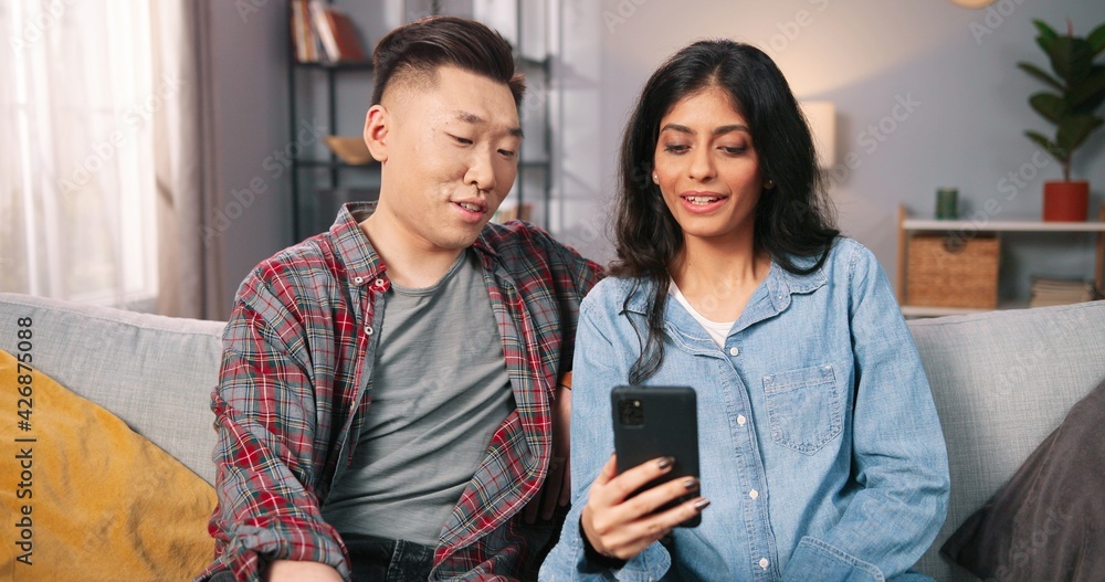 Portrait of multi-ethnic joyful happy couple wife and husband talking on video call online on smartphone while sitting on sofa in living room, videochatting, browsing on cellphone, leisure concept Portrait of multi-ethnic joyful happy couple wife and husband talking on video call online on smartphone while sitting on sofa in living room, videochatting, browsing on cellphone, leisure concept