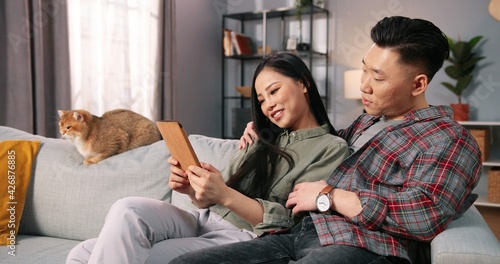 Portrait of Asian young happy lovely couple man and woman tapping online browsing on tablet device spending time at home together sitting on sofa and speaking discussing something on internet