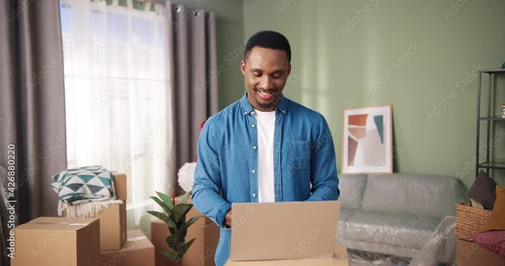 Portrait of African American joyful man standing in new apartment happy smiling and typing on laptop on carton box searching new furniture and decor for home. Relocation concept, relocating day Portrait of African American joyful man standing in new apartment happy smiling and typing on laptop on carton box searching new furniture and decor for home. Relocation concept, relocating day