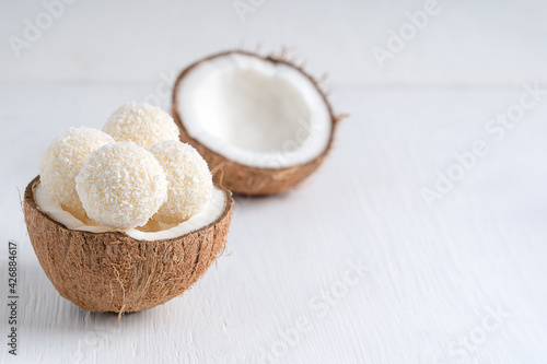 Coconut truffles or homemade organic energy balls with a filling of sweetened shredded coconut and curd cheese served in halved fresh coconut on white wooden table. Image with copy space, horizontal