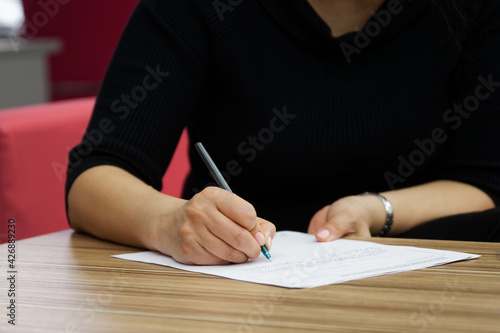 A woman in black clothes, sitting at a wooden table, fills out documents, a contract or a resume