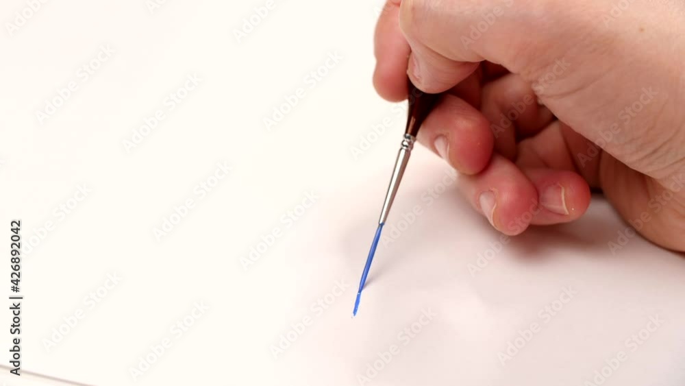 Artist's hand draws a thin blue line with watercolor or acrylic paint on canvas or white paper. Close-up shot of the female artist's hands painting using a paintbrush. Drawing concept Artist's hand draws a thin blue line with watercolor or acrylic paint on canvas or white paper. Close-up shot of the female artist's hands painting using a paintbrush. Drawing concept