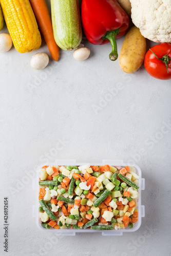 Rectangular plastic container with mix of frozen vegetables top view with copy space on gray background and fresh vegetables cauliflower, carrots, potatoes, mushrooms, zucchini, tomatoes, corn.