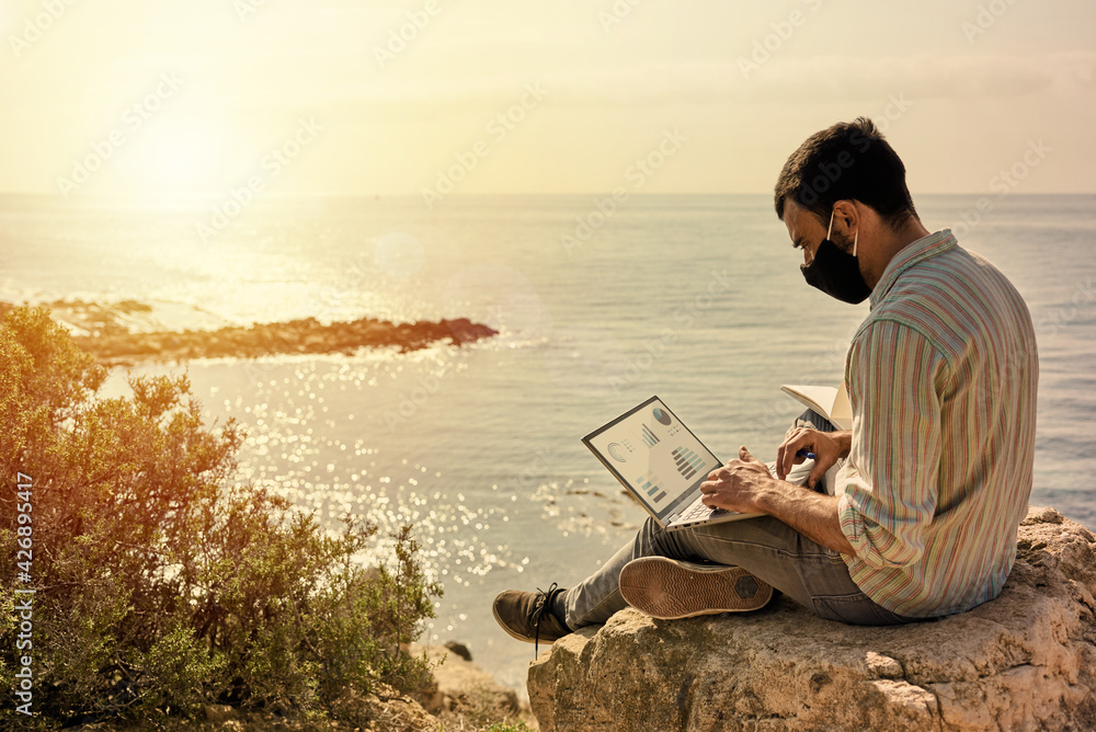 Man wears face mask while working remotely with his laptop from the beach