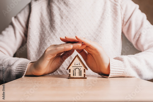 Woman holds and protects a wooden house with her hands with the sun on a light pink background. Sweet home