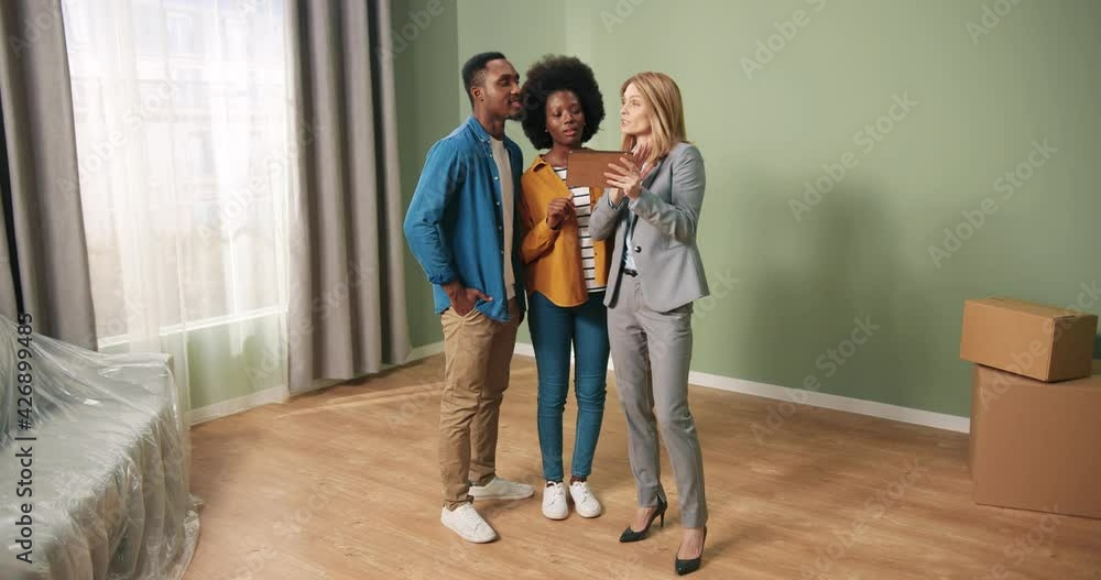 Caucasian joyful beautiful young real estate broker standing in apartment with African American couple, typing on tablet showing design of new apartment to clients. Realtor agent. Buying home concept Caucasian joyful beautiful young real estate broker standing in apartment with African American couple, typing on tablet showing design of new apartment to clients. Realtor agent. Buying home concept