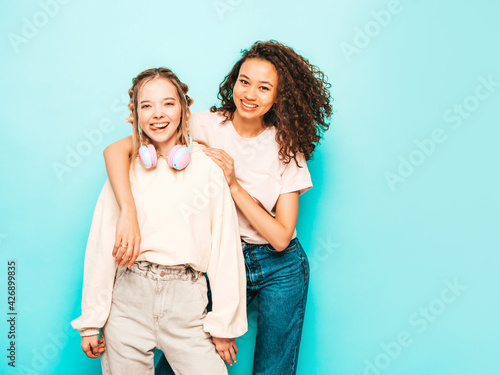 Two young beautiful smiling international hipster female in trendy summer clothes. Sexy carefree women posing near blue wall in studio. Positive models having fun. Concept of friendship © halayalex