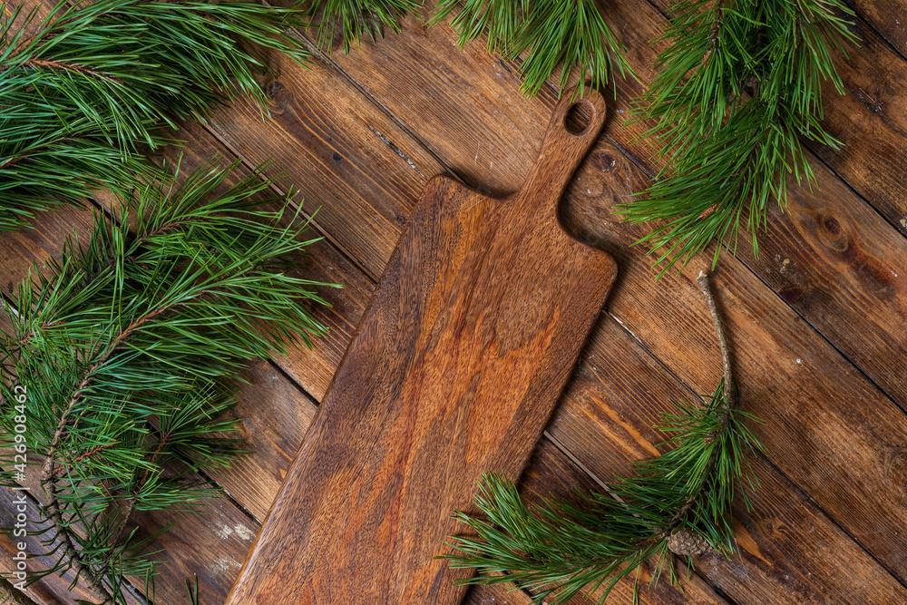 Cutting board on a wooden table background surrounded by pine branches, top view. Cutting board on a wooden table background surrounded by pine branches, top view.