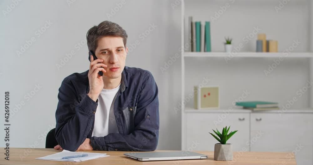 Serious caucasian millennial middle aged 30s businessman guy freelancer man sitting at table in office at home speaks on mobile phone conversation with client orders booking using wireless smartphone Serious caucasian millennial middle aged 30s businessman guy freelancer man sitting at table in office at home speaks on mobile phone conversation with client orders booking using wireless smartphone