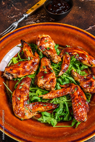 Baked chicken wings with sweet chili sauce on a plate with arugula. Dark background. Top view
