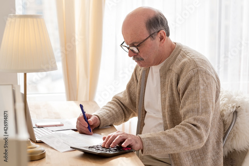 Senior man sitting with paperwork and using calculator while counting money