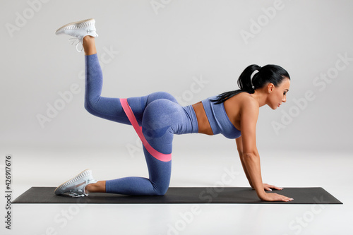 Fitness woman doing kickback exercise for glutes with resistance band on gray background. Athletic girl working out donkey kicks.