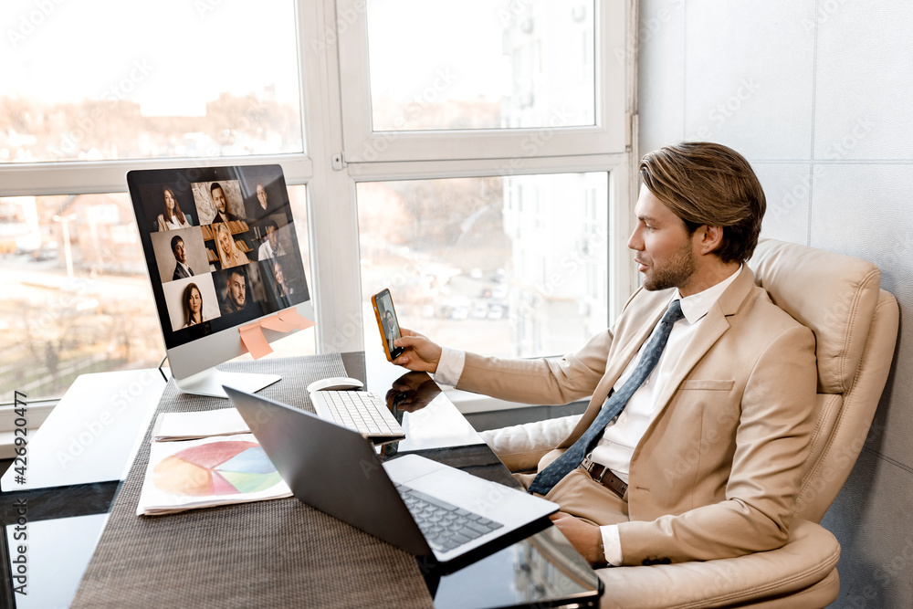 Stylish successful male boss is using computer and smartphone, having a video conference. Serious handsome businessman discussing with colleagues financial reports for project, remotely work concept Stylish successful male boss is using computer and smartphone, having a video conference. Serious handsome businessman discussing with colleagues financial reports for project, remotely work concept