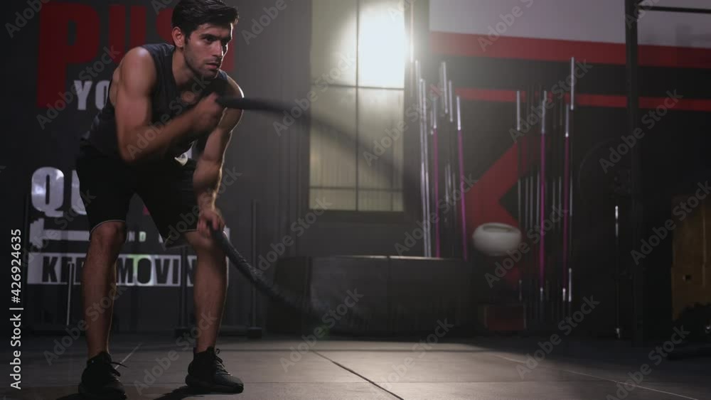 Athletic young man doing battle rope exercise in functional training fitness gym. Determined caucasian handsome man standing and doing cross fit exercise by battle rope. Healthcare and fitness in gym.