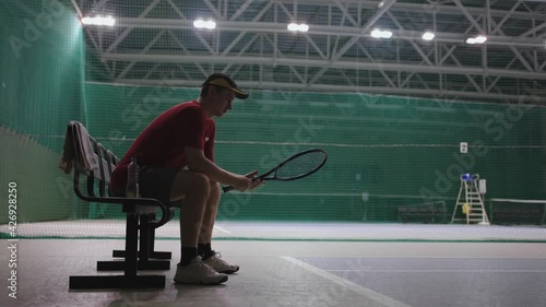 male tennis player is sitting on bench in tennis court after training or match, resiting and twisting racquet in hand photo
