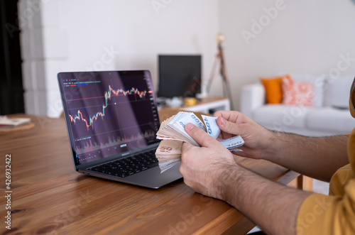 man counting his cash money while watching money and stock market graphs from leptop at his home.