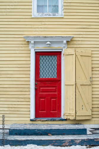 The exterior wall of a yellow wood clapboard heritage house. The door of the building is bright red with a small glass window. There's a lace curtain in the double hung window that has white wood trim © Dolores  Harvey