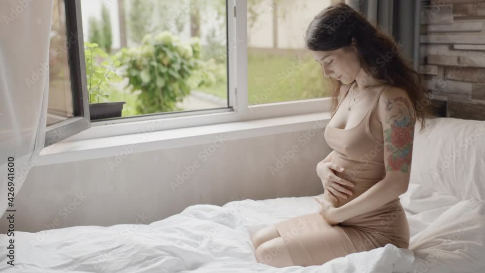Pretty expectant mother sitting in light room. Happy pregnant woman stroking belly near window indoors. Future mom petting her stomach. Pregnant woman smiling to unborn baby Pretty expectant mother sitting in light room. Happy pregnant woman stroking belly near window indoors. Future mom petting her stomach. Pregnant woman smiling to unborn baby
