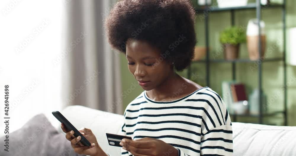 Close up of young beautiful african american woman sitting at home on sofa holding smartphone and bank card doing online shopping. Close up of young beautiful african american woman sitting at home on sofa holding smartphone and bank card doing online shopping.