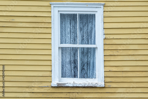 The exterior of a vintage yellow coloured wall with narrow wood cape cod clapboard siding. In the center of the house  there s a closed double hung window with a white lace curtain. 