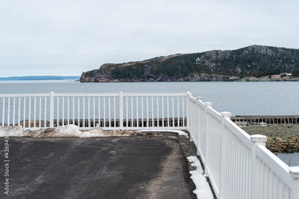 Obraz premium A long white metal fence and corner join with post caps along the edge of a paved parking lot overlooking the blue ocean. There's a tree covered island in the background under white cloudy skies. 
