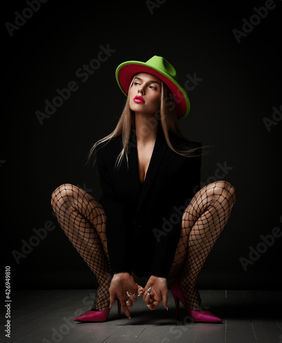 Naughty arrogant young sexy woman in jacket, fishnet tights stockings, pink high-heeled shoes and hat sits squatted looking at upper corner over dark background. Sexual games, bdsm, glamour concept