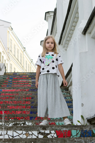 A stylish model, a six-seven-year-old girl with blond hair, stands on a bright stone staircase in a city street. © bela_zamsha