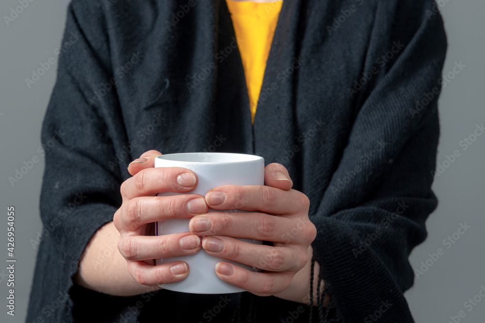 woman in a black cloak is holding white mug with hot drink in both hands woman in a black cloak is holding white mug with hot drink in both hands