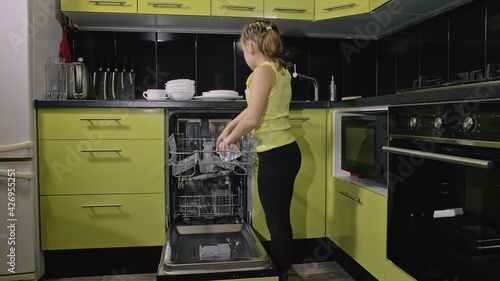 Smart girl learning to use dishwasher. Stylish modern Built In Kitchen Appliances in green black. Child is putting clean dishes.