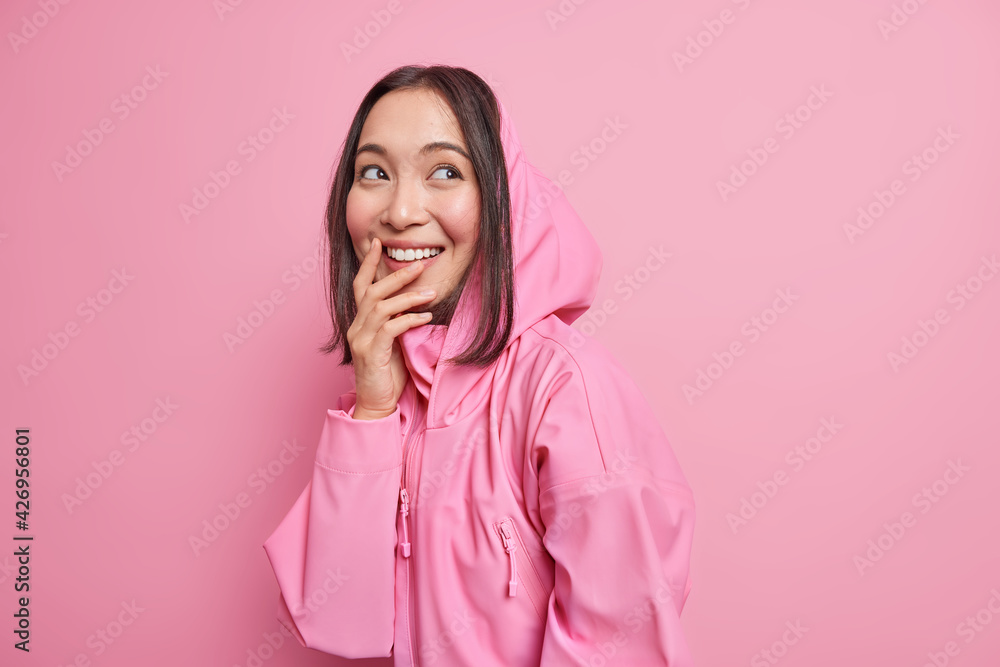 Cheerful Asian female teenager gazes aside daydreams and remembers lovely moment smiles broadly dressed in hoodie expresses positive candid emotions isolated over pink background. Youth lifestyle Cheerful Asian female teenager gazes aside daydreams and remembers lovely moment smiles broadly dressed in hoodie expresses positive candid emotions isolated over pink background. Youth lifestyle