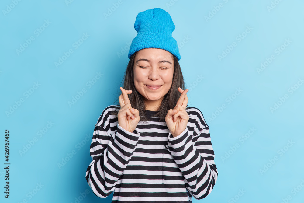 Happy pleased Asian teenage girl crosses fingers anticipates for results keeps eyes shut believes dreams come true wears casual striped jumper and hat isolated over blue background. Good luck Happy pleased Asian teenage girl crosses fingers anticipates for results keeps eyes shut believes dreams come true wears casual striped jumper and hat isolated over blue background. Good luck