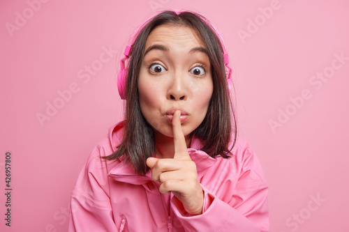Tableau sur toile Surprised Asian woman looks with mysterious expression tells to be quiet makes taboo gesture listens music via wireless headphones wears jacket isolated over pink background