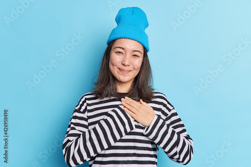Happy beautiful Asian woman makes gratitude gesture presses palms to heart feels touched pleased by compliments says thank you expresses thankfullness wears blue hat striped jumper poses indoor