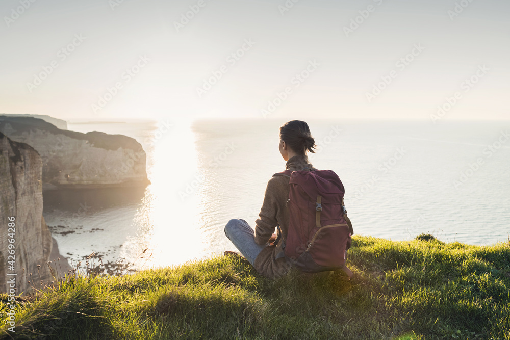 Young woman traveler with backpack sitting and looking at sunset above a sea over beautiful cliffs background, Travel, active lifestyle and summer holiday concept	