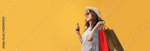 Beautiful Asian shopaholic woman smiles and happy while carrying shopping bags with smartphones and credit card on yellow background. Summer sale, Mid-year sales banner concept.