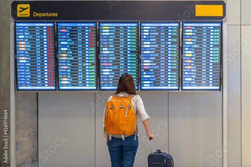 Back side of asian woman backpacker or traveler with luggage standing over the flight board for check-in at the flight information screen in modern an airport, travel and transportation concept