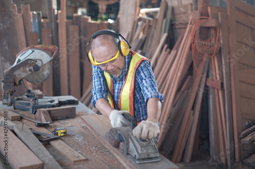 An old Asian male carpenter is working on an electric planer in a wood factory There are also other tools in the craft manufacturing industry, such as tape measure, electric saw, etc.