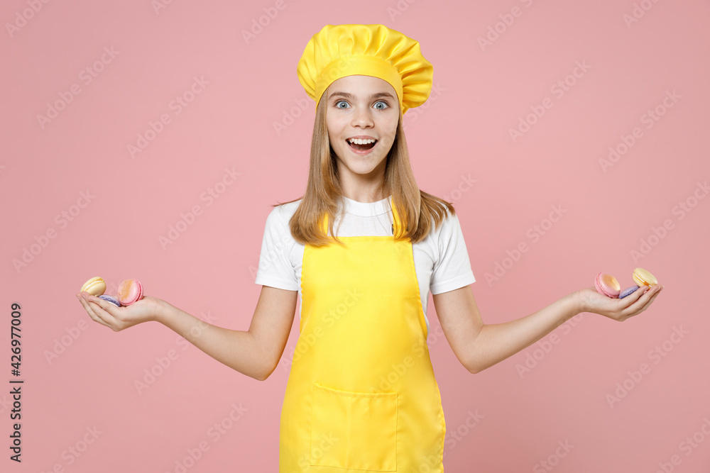 Teen surprised happy girl chef cook confectioner baker housewife in yellow apron white t-shirt toque cap holding macaroons in outstretched hands isolated on pastel pink background Food cake concept. Teen surprised happy girl chef cook confectioner baker housewife in yellow apron white t-shirt toque cap holding macaroons in outstretched hands isolated on pastel pink background Food cake concept.