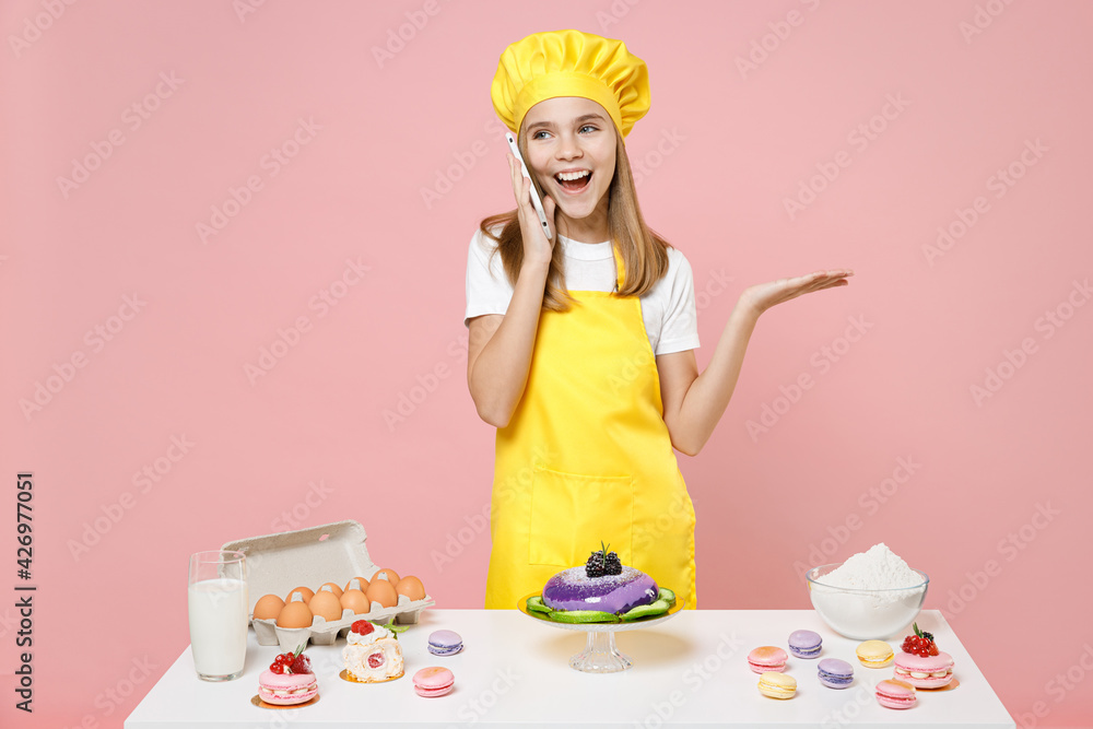 Teen girl chef cook confectioner baker mother's helper in yellow apron cap at table talk by mobile phone isolated on pastel pink background studio. Mousse cake cooking food process workshop concept. Teen girl chef cook confectioner baker mother's helper in yellow apron cap at table talk by mobile phone isolated on pastel pink background studio. Mousse cake cooking food process workshop concept.
